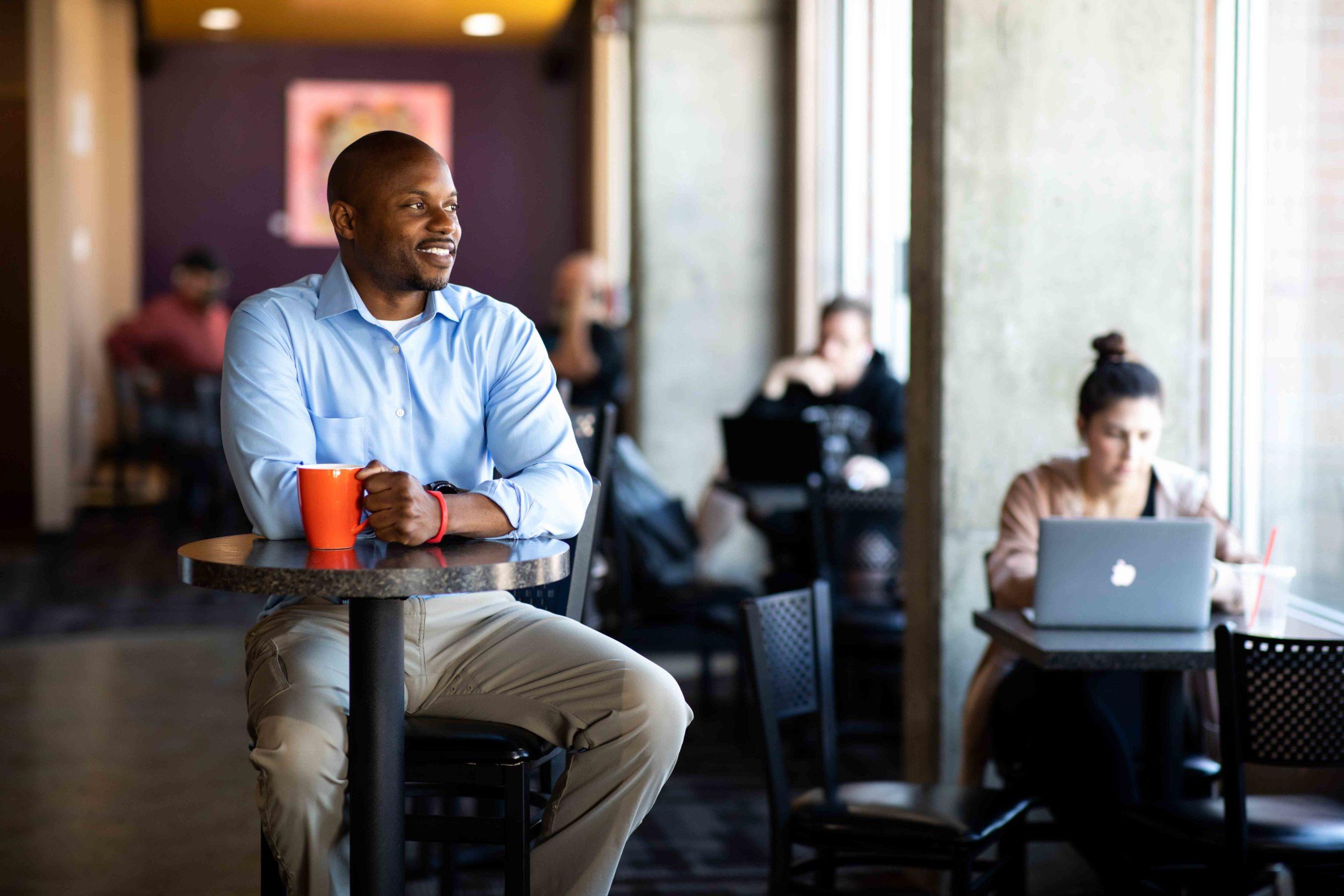 MSU Denver Grad Ryan Cobbins sitting in a coffee shop with a mug in his hand, looking out the window.