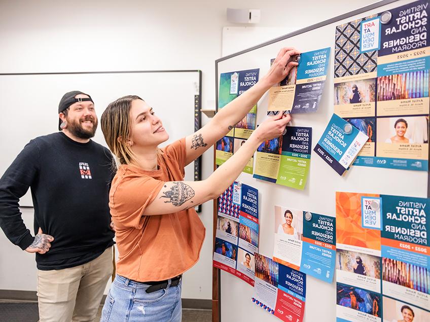 Students post finalized examples on a white board, for their project which creates advertisements for the Visiting Artist Scholar and Designer Program. In use are many bright and complementary color palettes including teal, sky blue, orange, lime green and navy, along with a dark cream and light lavender. Both students happen to have prominent tattoos showing on their arms.
