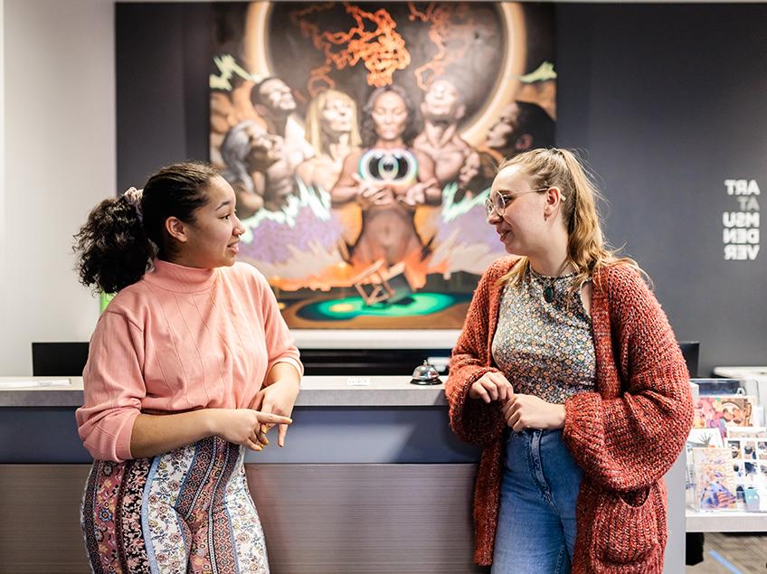 Two students chat while standing at the front desk of the Art Deparment. Behind them is a painting of a diverse group of individuals representing the many peoples of earth. They hold a central floating orb and cube, in poses of reflection and contemplation.