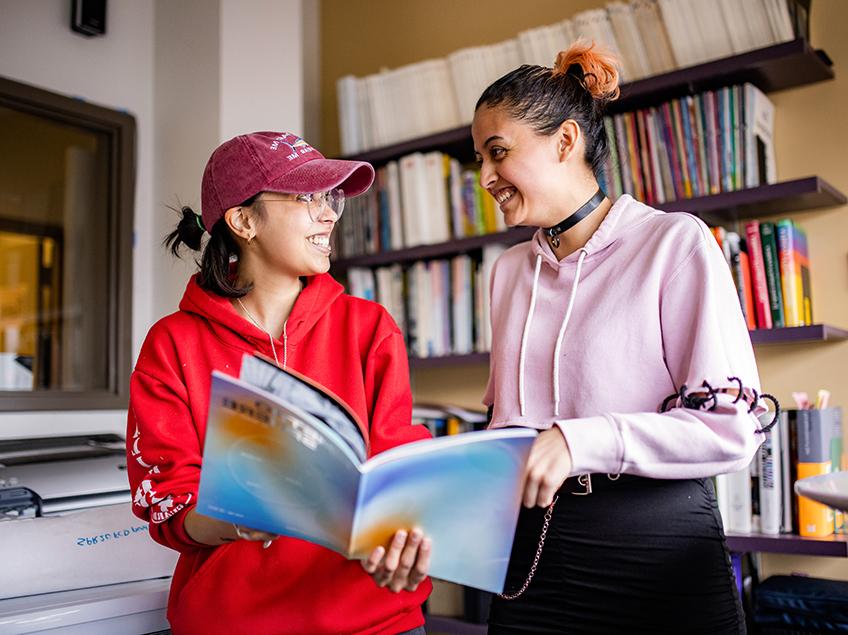 Two Communication Design students flip through the Metrosphere magazine, which they helped to design and create. They stand in the CDES library in front of the many books and periodicals which are available for student research and reading.