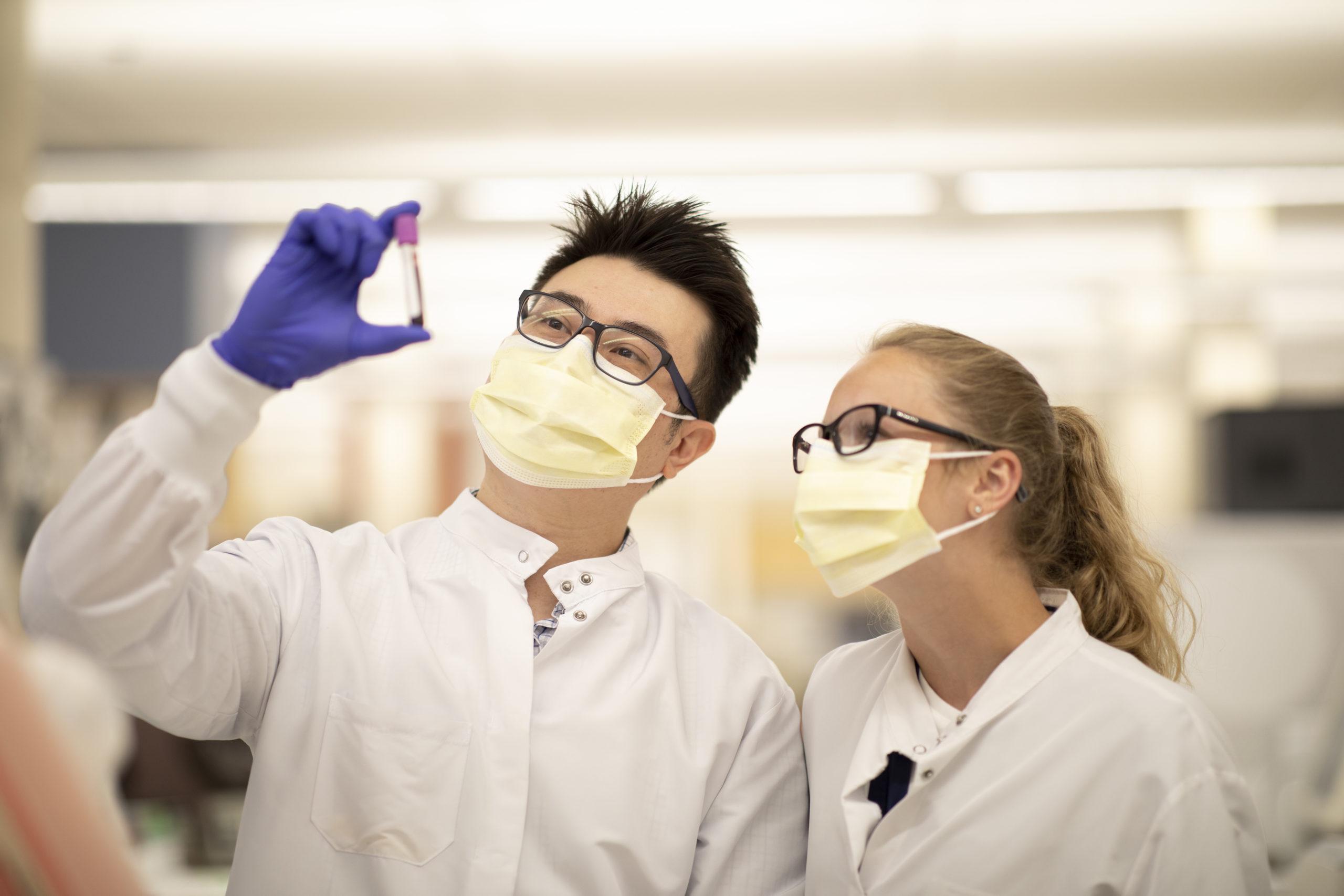 Two people in lab coats and masks examining a test tube sample.