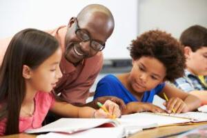 An African-American male teacher works with two Elementary school children at a desk.