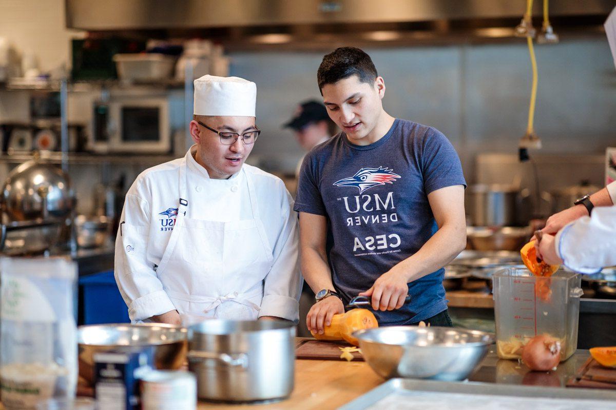 一名身穿蓝色衬衫的学生正在切蔬菜，旁边是一名身穿白色厨师服的学生