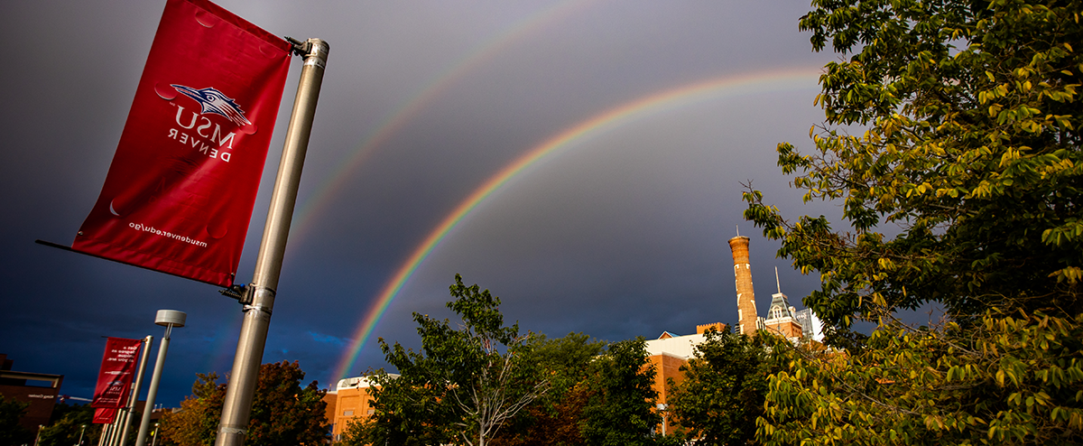 Photograph of a double rainbow above the Tivoli building with a red MSU Denver flag in the foreground.